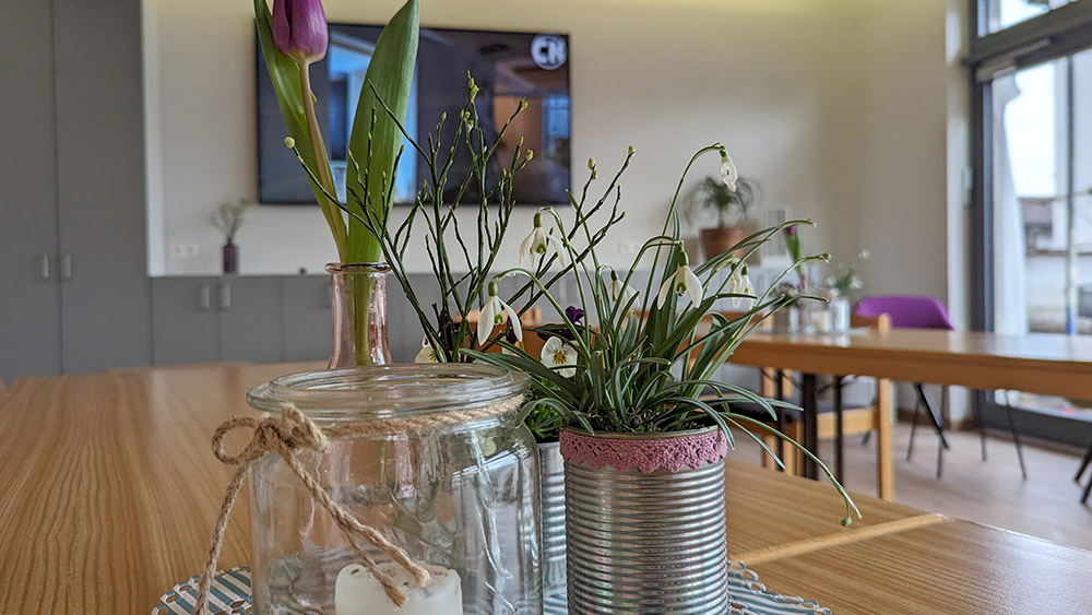 Frauencafé Foto eines Raums. Im Vordergrund scharf zu stehen steht mittig eine Tischdekoration mit Blumen und Kerzen. Im Hintergrund sind ein Tisch und ein großer Fernseher zu sehen.